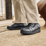 Ein Mann in beiger Hose und schwarzen New Feet Herrenstiefeln steht auf grauem Untergrund